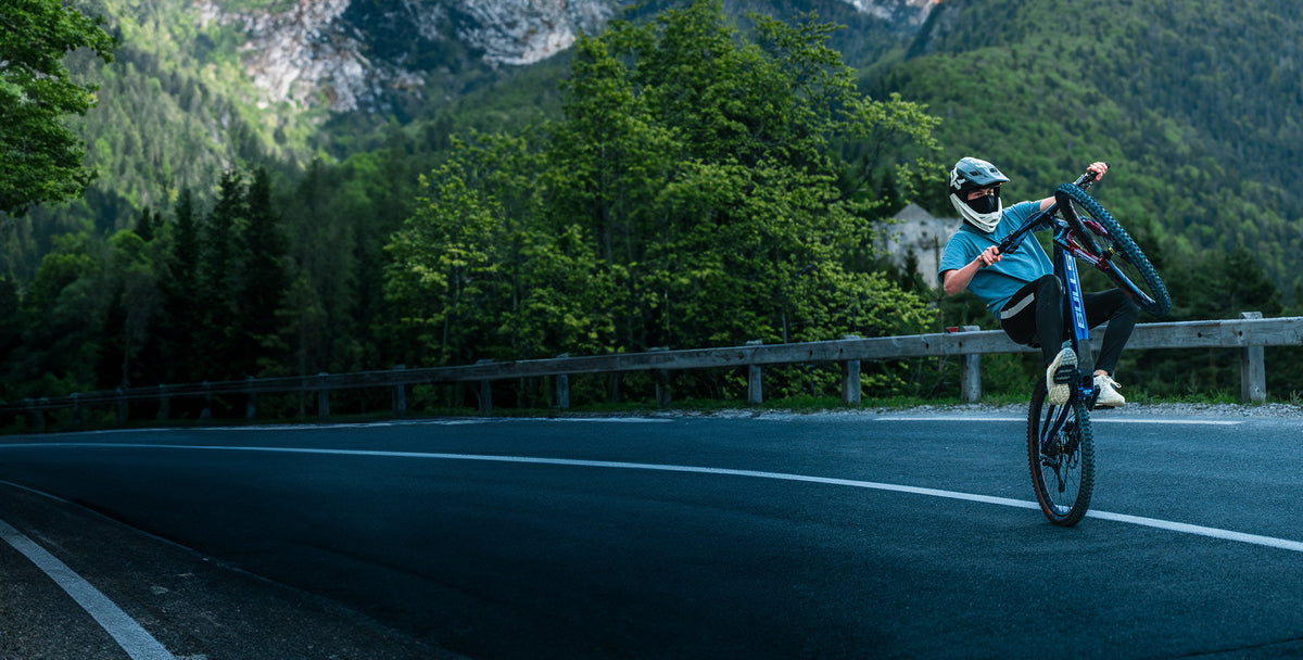 Niko von NoHandMTB zieht auf einer Bergstraße einen Wheelie auf dem Mountainbike, im Fullface-Helm mit Maske